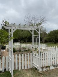 Three foot high white picket fence around a garden with a white trellis entrance. In the background you can see the white picket shelves a friend's dad originally built for a bedroom.