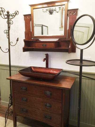 Bronze glass sink on top of an antique 3-drawer dresser. An antique mirror hangs on the wall. On the left is  bronze coat rack stand for towels. To the right of the sink is a free-standing adjustable mirror with hand-towel bar.