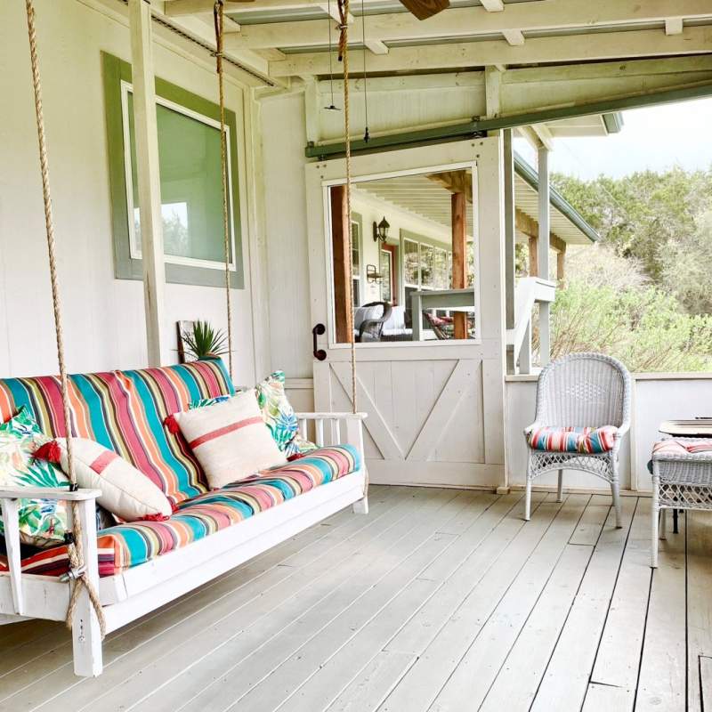A large futon porch swing hangs on a huge screened porch. Two white wicker side chairs frame the corner seating area. 