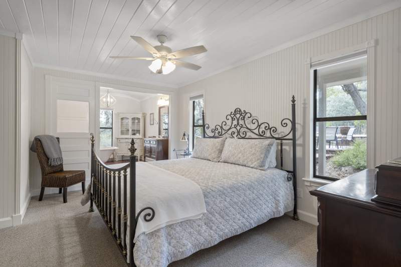 Light, spacious bedroom with king size bed and a side chair. In the background is an elegant bathroom, slightly elevated with wood flooring. It has a chandelier over a footed soaking tub, and a white antique cabinet in the corner. The sink is in a wood stained antique dresser with a mirror hung above it.