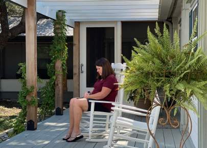 A woman relaxing in a white rocking chair under the pergola of the front porch.