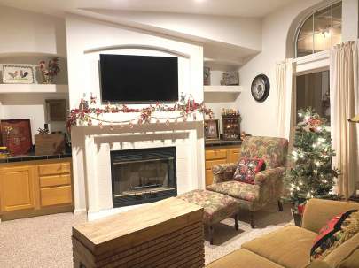 Living area with a flat screen TV over a fireplace. The mantel of garland has Santa figurines. A small tree stands between a chair and sofa.