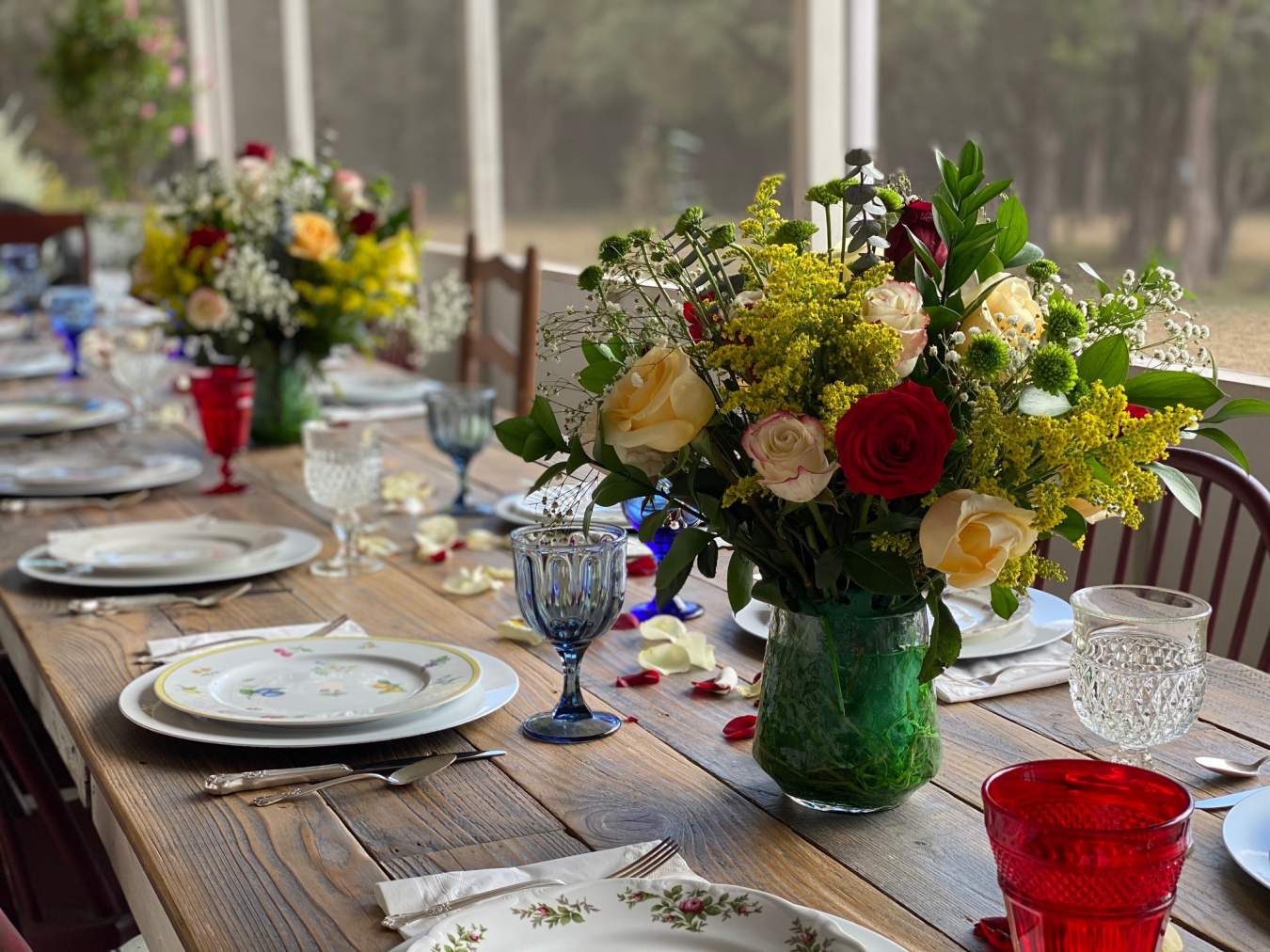 A beautifully appointed farmhouse table set with floral China plates, colorful vintage goblets and two bouquets of roses.