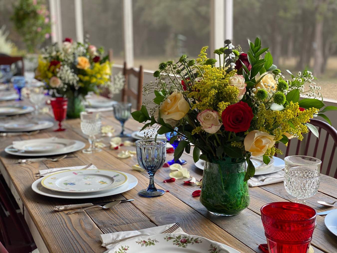Breakfast on the screened porch A beautifully appointed farmhouse table set with floral China plates, colorful vintage goblets and two bouquets of roses.