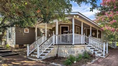 BellaVida Hause, a farmhouse style short-term rental. Charming house with three sets of steps up to the porch, each with white handrails and newel posts topped with round finials. The house is surrounded by oaks and pink blooming crepe myrtle trees.