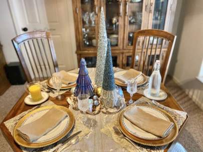 Table set for four. The centerpiece is three different heights of light blue, gold and pearl Christmas trees encircled by ribbon and glass ornaments.