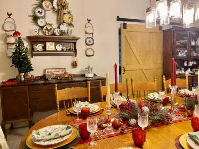 Colorful Christmas table setting with a centerpiece of red and white candles, bells and greenery. The buffet in the background has a small Christmas tree and other decor on it.
