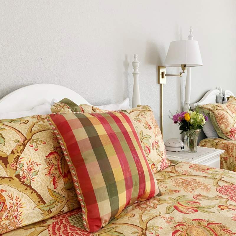 A close-up of two beds with a plush floral design comforter and shams with a coordinating plaid decorative pillow in front. A night stand between the beds has a vase of fresh flowers and a wall lamp hangs on the wall above it.