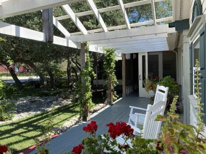 Two white rocking chairs on a porch with sun shining through a pergola above. Red geraniums are in the foreground.