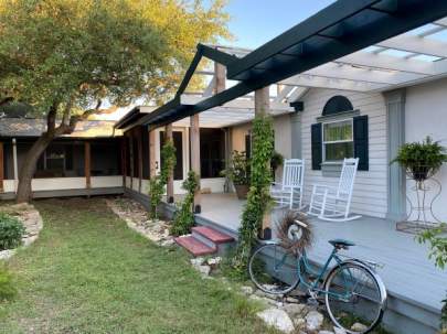 Elevated porch with a pergola and vines growing up the support beams. Two inviting white rockers with plant stands on each side. A vintage bike leans against the porch. The far end of the porch is covered and screened.