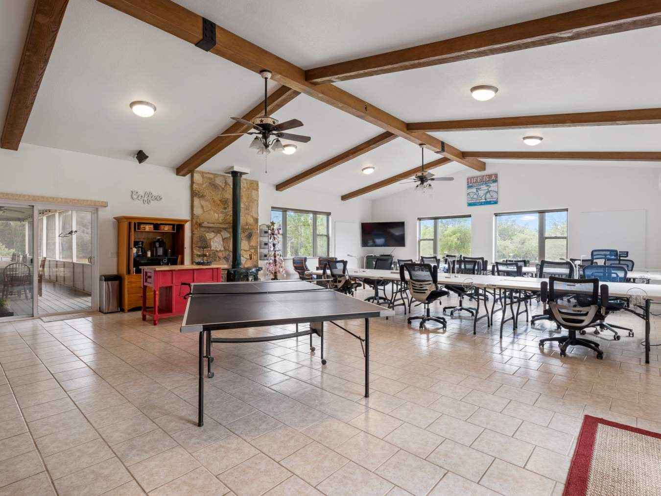 Large room with a ping pong table in the foreground. Beyond are several 6-foot tables with adjustable rolling chairs and a large flat screen TV.