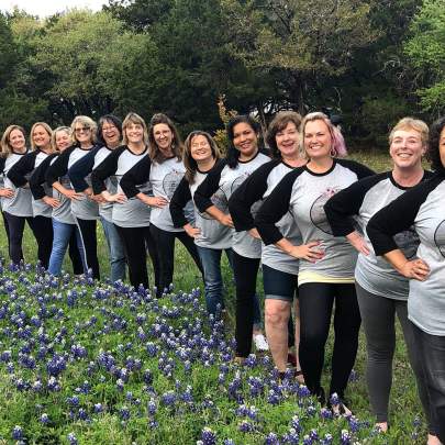 A line-up of thirteen ladies wearing matching shirts, standing in a field of bluebonnets at BellaVida Retreats.