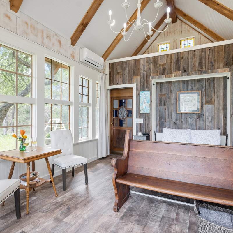 Light, airy chapel bedroom. Vintage windows surround the room with views of the scenic landscape. A queen bed with old church pew at the foot. A small table and two white side chairs.