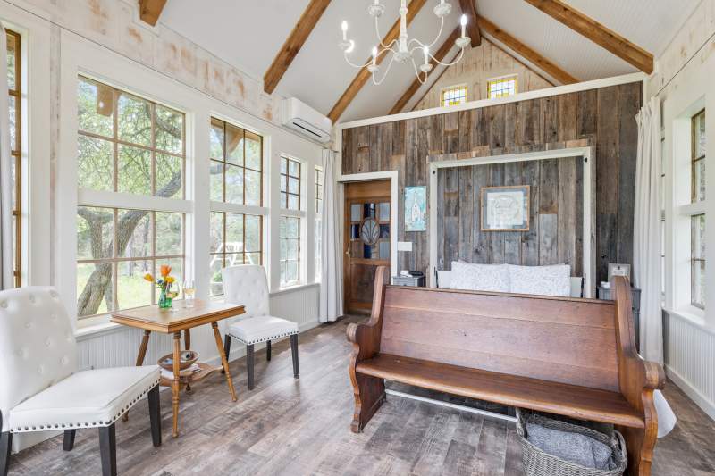 Light-filled room surrounded with multi-pane windows and cathedral ceiling. Drapes are open and gathered at the wall corners. A chandelier hangs in the center. Ship lap walls and wood grain flooring offer the feel of a country church. An old oak church pew is at the foot of the queen bed. Along the side wall are two white upholstered chairs with a small wood table between them with a vase of fresh flowers.