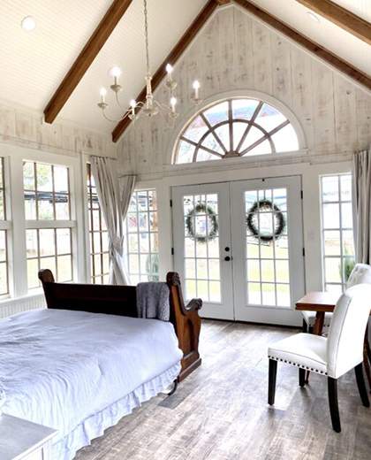 Light-filled room surrounded with multi-pane windows and cathedral ceiling. Drapes are open and gathered at the wall corners. A chandelier hangs in the center. Ship lap walls and wood grain flooring offer the feel of a country church. Double French doors with a large radius window are in the background. An old oak church pew is at the foot of the queen bed with a gray comforter cover. Along the side wall are two white upholstered chairs with a small wood table between them.