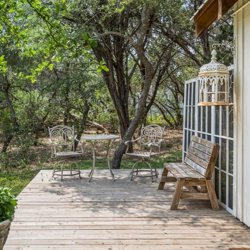 Private patio with wood decking framed by trees and a French door style screen wall. In the shade of the trees is an ornate bistro set for two. Against the screen wall is a wood bench and hanging next to it a large cathedral style bird cage with twinkle lights in it. 