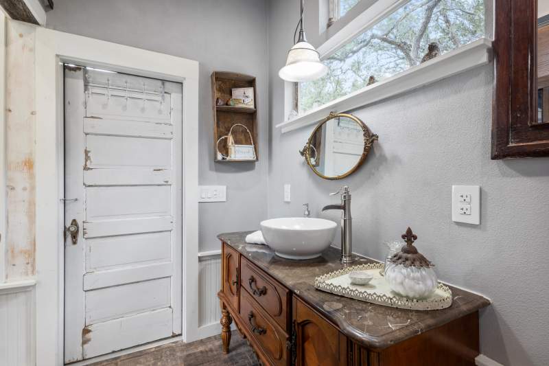A top mounted white bowl sinks sits on a granite top vanity with ornate stained cabinetry. An antique gold rimmed mirror hangs above the vanity with a horizontal window inviting nature inside. To the left is a worn, vintage farm-house-style door that leads outside to a private porch.