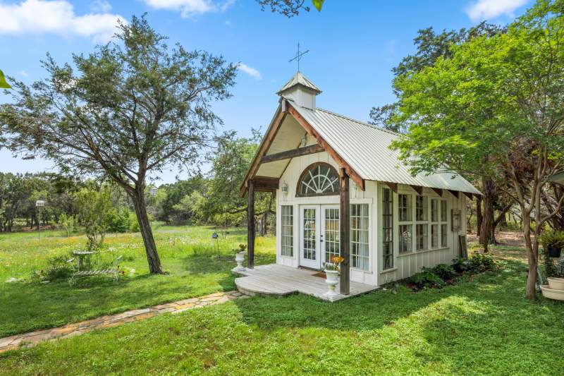 A small chapel sits among trees, green grass and flowers. A rock walkway leads to the entry. The church structure is of vintage windows and French-style front doors. Above the doors is a large arched vintage window. The front portion of the roof has a small cupola with a narrow metal cross on it.