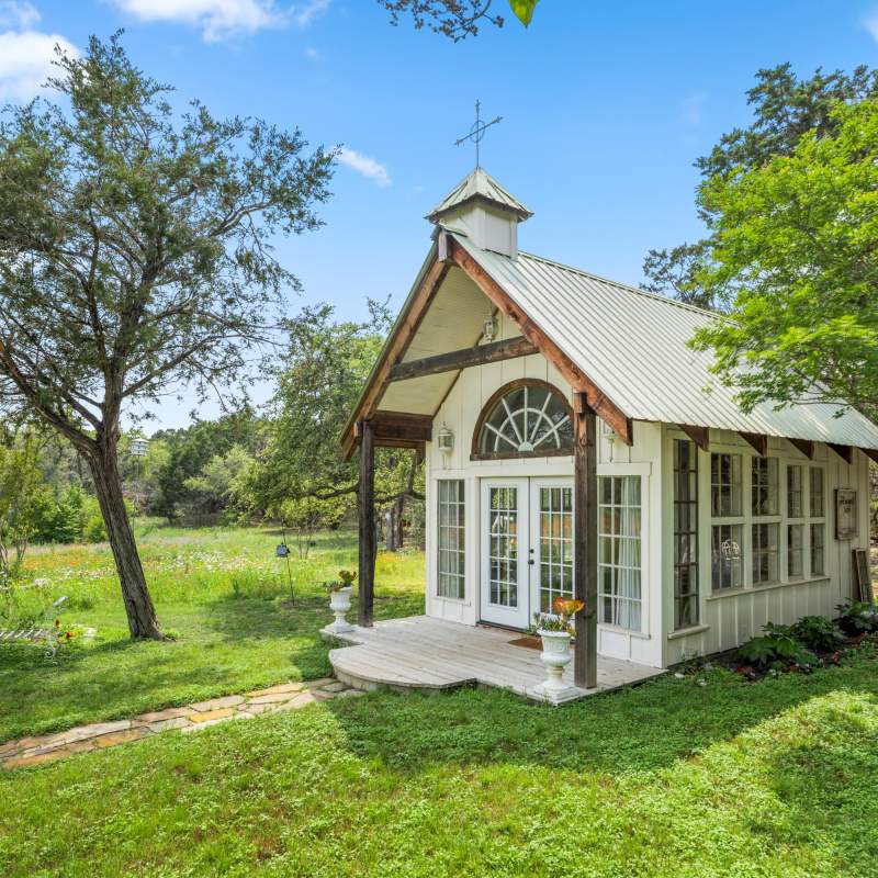 Trees frame a white wedding chapel with vintage windows and French front doors. Above the doors is a vintage arch window. On the metal roof is cupola with a cross. A stone walkway leads to the front entry. 