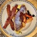 Peach croissant French toast with powdered sugar sprinkle topped with peach slices and blueberries. Plus a side of two slices of bacon served on a formal China plate.