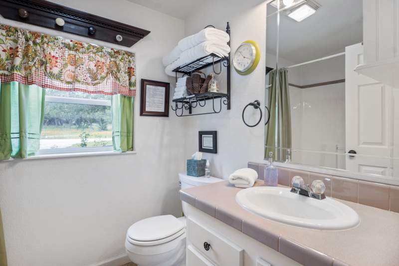 The two rooms with twin beds share a bathroom in the hallway between them. It has colorful curtains on the one window, a tub with shower enclosure and a counter with a sink. Above the toilet is a black wrought iron shelf with folded towels.