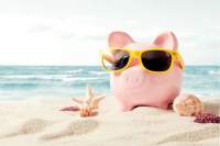 Pink piggy bank wearing sunglasses sitting on a sandy ocean beach with a starfish and sea shell.
