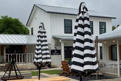 White modern farmhouse style two-story building with black trim. Outdoor social seating area with a fire pit, picnic tables with black and white stripe umbrellas for shade.
