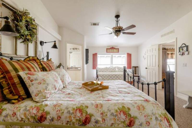 Light, airy bedroom with floral queen bedding. Farm style adjustable lamps attach from the wall on each side of the bed. In the background is a twin size day bed against a window. A door on the left opens to a bathroom. The entry door is open on the right side.