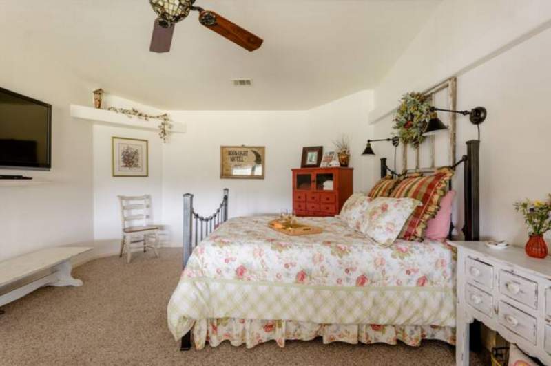 Black wrought-iron queen bed with two, black farmhouse style, wall-mounted reading lamps above the headboard. The opposite wall from the bed is a wall-mounted flat screen TV and below it, a white low table for luggage. There is a white side chair on an angled wall and a red tall chest with glass doors.