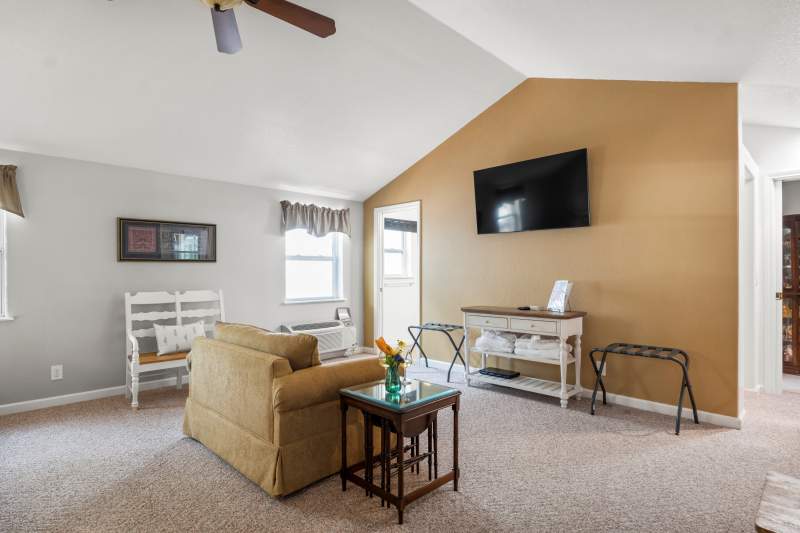 Sitting area with flat screen TV mounted on the wall. Underneath is a farm style cabinet with a lamp and DVD player. A luggage rack it on the left. In the foreground are two comfortable upholstered chairs with a small table in between.