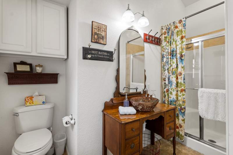 Bright lighting illuminates an antique dresser repurposed into a bathroom vanity with a glass surface mount sink. There is a step-in shower and white bath mat and hand towel.