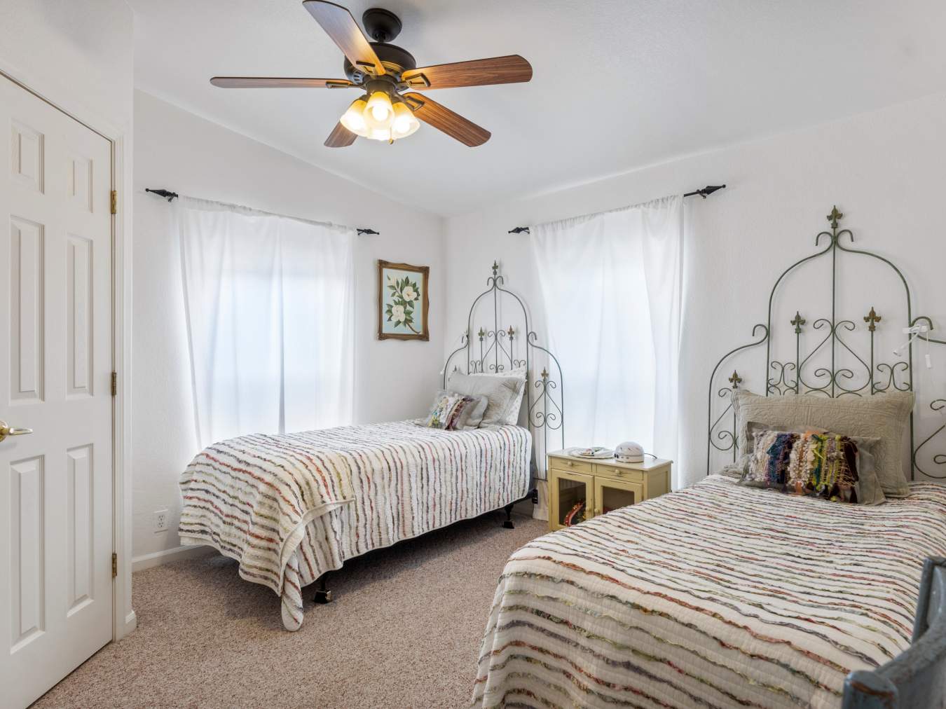 Light and airy bedroom with two twin beds, each with a colorful striped quilt and pale green iron trellis headboard. The room has two windows with white drapes and a ceiling fan.