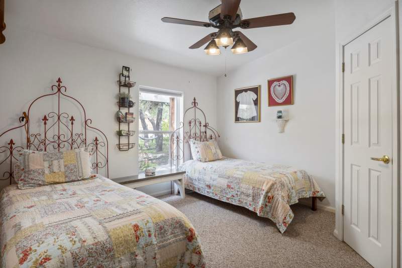 Two twin beds each with matching patchwork quilts and pillow shams. The headboards are red iron garden trellises. An antique baby dress is framed on the wall and a framed crocheted heart, There is one window and a ceiling fan.