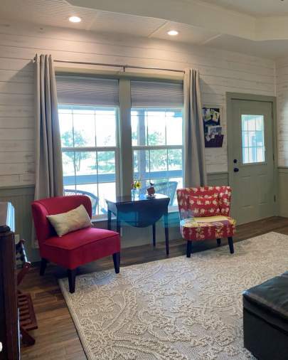 Sitting area with two red upholstered chairs on each side of a small drop leaf table. The wood grain floor has a large area rug. Light is coming through the window behind the table. There's a exterior entry door with a private porch outside.