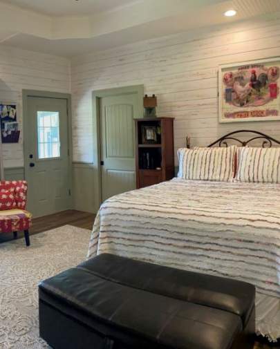 Bedroom with white ship lap walls., queen bed, and a large ottoman at the foot of the bed. There are two upholstered side chairs and a pocket door for the bathroom.