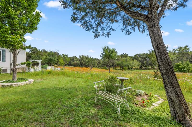 Spring time view from Howard Chapel. In the foreground is a pale green wrought iron vintage bench in front of a small flower bed with a bird bath. In the distance is white picket fencing around a garden and  a large field of colorful Texas wildflowers.