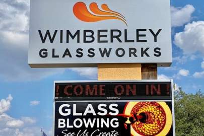 Wimberley Glassworks Signage with logo of flame. Below it is an electronic sign inviting you to stop in and see their glassblowing demos.