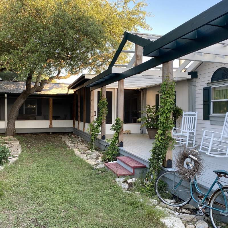 Long, elevated front porch with vines growing up the supports and onto the pergola. Two white rockers and plants adorn the porch and a vintage bike leans up against it. 