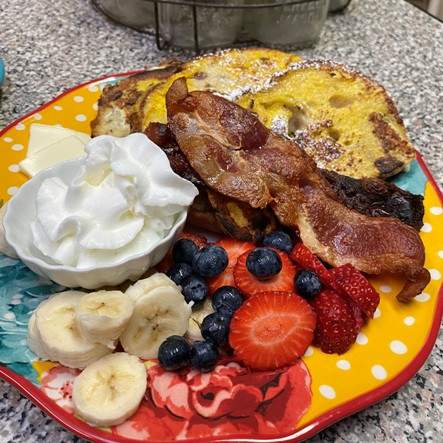 Colorful ceramic breakfast plate and on it, French toast, crispy bacon, side of fruit with strawberries, blueberries and bananas with fresh, whipped cream. Yum!