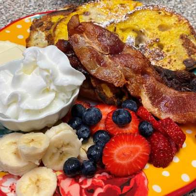 A breakfast plate of French toast, crispy bacon, side of fruit with strawberries, blueberries and bananas with fresh, whipped cream. Yum!