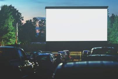 It's dusk at a drive-in movie theatre with the huge movie screen in the background and in the foreground many cars parked with occupants watching a movie.