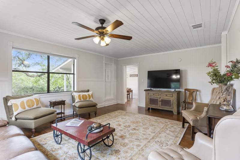 Spacious living room with stained wood floors,an area rug and white ship lap walls and ceiling. There is a brown leather sofa sleeper, table for games with two side chairs, two upholstered chairs and a TV cabinet with a large flat screen TV. It's a light airy room and through the double windows you can view the green trees and landscape outside.