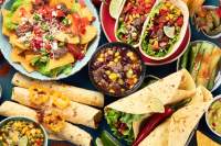 Colorful table of Mexican dishes of black beans, tacos, nachos with sliced avocado, and taquitas. 