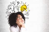 Woman with hands at her chin looking upward with a questioning look. A whiteboard behind her has a yellow light bulb with question marks surrounding it.