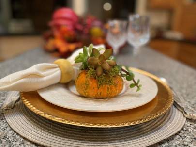 Miniature pumpkin with small succulent arrangement on top as a table arrangement at each place setting. Behind the pumpkin is a rolled, cream-colored napkin with a gold color napkin ring. It's on a China plate,  topped on a gold metallic charger.