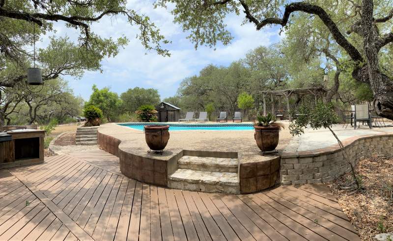 Foreground are two steps leading up to a swimming pool. In the background is a small house or shed structure and a cabana. Just left of the steps is a rustic outdoor kitchen.