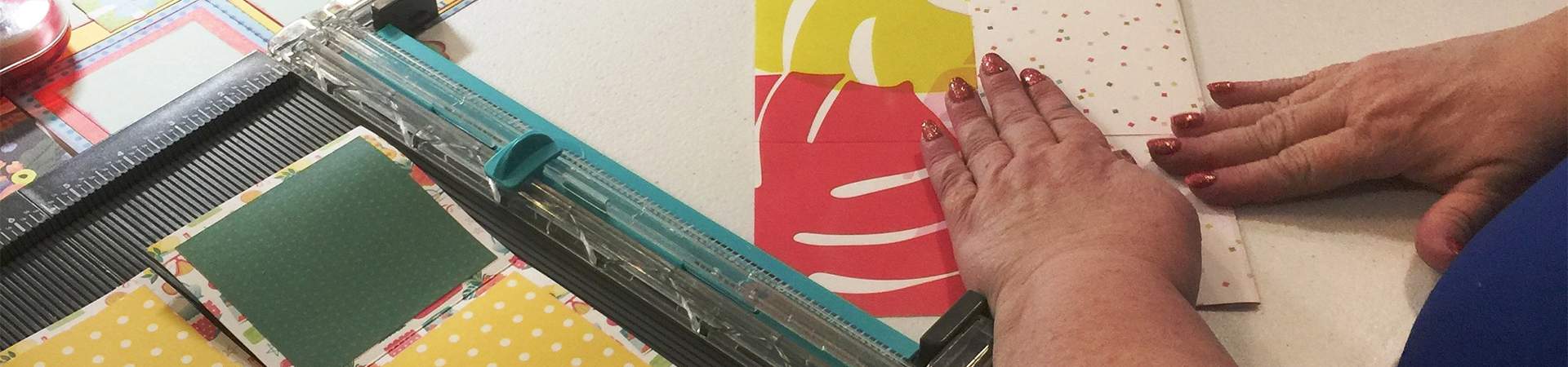 Only the arms and hands of a woman are visible as she folds colorful paper  for a paper crafting project. On her left is a paper cutter with part of the project laid out.