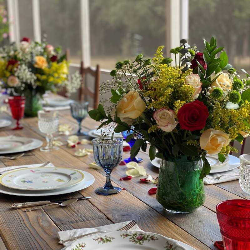 Farmhouse style table with mixed patterns of table China. Crystal glassware is of mixed colors: red, clear, gray and cobalt. The table is adorned with lush floral centerpieces of colorful roses and baby's breath.